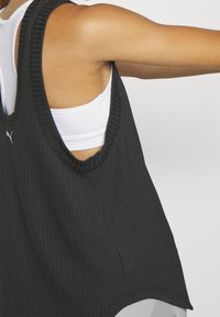 Czarny prążkowany top na ramiączkach z szerokimi otworami na ramiona i okrągłym dekoltem, z białym logo z przodu, noszony na białym biustonoszu sportowym.