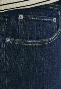 Záber na vrecko tmavomodrých riflí s oranžovým prešitím a kovovými nitmi, doplnené o lem bieleho a čierno pruhovaného trička.