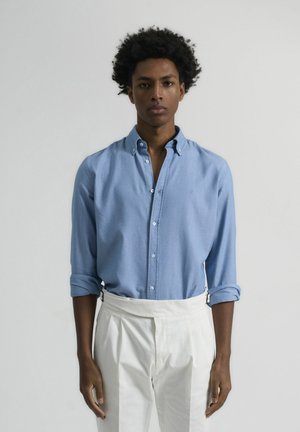 Camisa de botón azul claro con una textura suave, mangas enrolladas y un pequeño logo en el pecho, combinada con pantalones blancos de cintura alta.