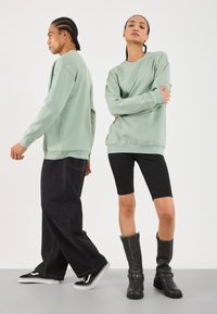 Dos modelos que llevan sudaderas verde claro; uno con jeans anchos negros y zapatillas, y el otro con pantalones cortos negros y botas negras con hebilla, contra un fondo blanco.