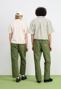 Deux individus se tiennent dos à dos, portant une chemise beige rayée et une chemise gris clair. Tous deux portent un pantalon cargo vert avec des poches et des étiquettes.
