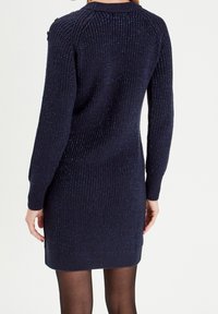 Robe-pull en maille bleu marine avec des paillettes métalliques, texture côtelée, manches longues, silhouette ajustée et encolure arrondie.