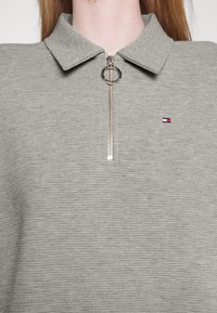 Pulóver gris de media cremallera con una textura acanalada, que presenta un cuello clásico y un tirador de cremallera circular, acentuado por un pequeño logotipo.