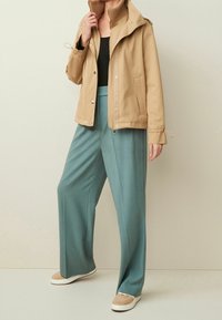 Beige waterproof jacket with a high collar and adjustable cuffs, paired with teal wide-leg trousers and beige slip-on sneakers.