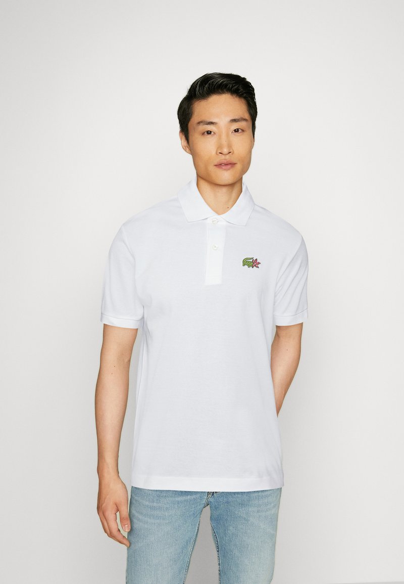 Polo shirt bianco con colletto classico, maniche corte e un piccolo logo ricamato raffigurante un coccodrillo e un fiore sul lato sinistro del petto.