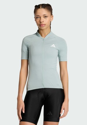 Młoda kobieta w jasnoszarej, krótkorękawowej koszulce kolarskiej i czarnych, wyściełanych spodenkach kolarskich, stojąca na jednolitym tle.