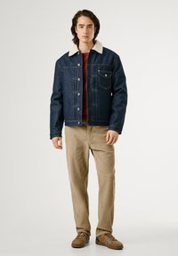 Chaqueta de mezclilla con cuello de borreguillo, color azul oscuro, bolsillo delantero con parche, combinada con pantalones de pana color marrón claro y zapatillas marrones.