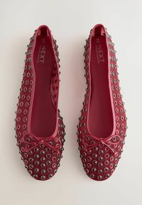 Ballerines rouges avec un extérieur lisse, ornées d'œillets argentés et noirs, présentant un détail de nœud central au niveau des orteils.