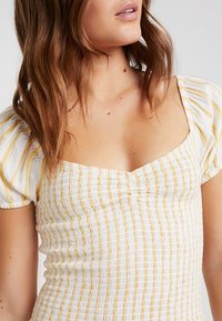 Biało-żółty top w paski z bufkami na rękawach i kwadratowym dekoltem. Tkanina ma marszczoną, ściągaczową fakturę na całej powierzchni.
