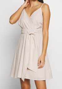 Kvinna i en ärmlös beige omlottklänning med en knuten midjebälte, stående mot en ljus, enfärgad bakgrund.