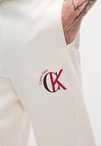 Białe spodnie dresowe o gładkiej fakturze; z czerwonym logo Calvin Klein po lewej stronie. Projekt zawiera detal w postaci bocznej kieszeni.