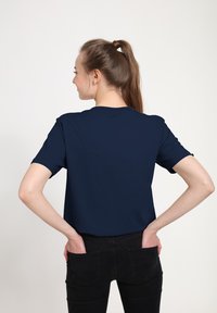 Námořnická modrá krátká rukávová trička s kulatým výstřihem, vyrobené z hladkého materiálu, nošené s černými přiléhavými džíny. Viditelný design na zádech, minimální detaily.