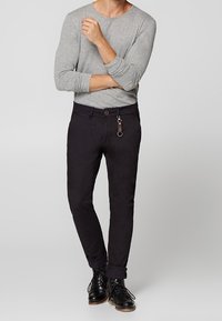 Pantaloni neri slim fit realizzati in cotone, con chiusura a bottone e dettaglio anello per chiavi. Indossati con una camicia grigia a maniche lunghe.
