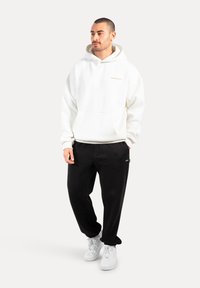 Uomo in piedi che indossa una felpa bianca con cappuccio, pantaloni neri e sneakers bianche su uno sfondo chiaro uniforme.