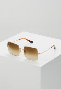 Ochelari de soare din metal aurit, cu lentile pătrate, având un ton de gradient maroniu și brațe din acetat cu model țestoasă, expuși pe un piedestal alb.