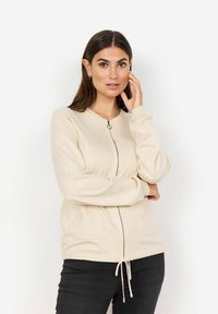 Cardigan beige con zip frontale, scollo rotondo, polsini a coste, due tasche frontali e cordino regolabile all'orlo.