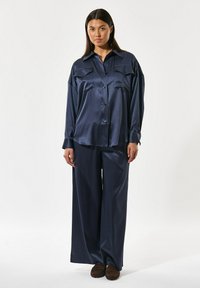 Mørkeblå satin loungewear sæt med en langærmet skjorte med knaplukning og to brystlommer samt bukser med vide ben. Blød, glat tekstur.