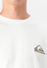 Moški vrat in zgornji del prsnega koša, oblečen v belo majico z okroglim izrezom in majhnim sivim logotipom gorske valove na levi strani.