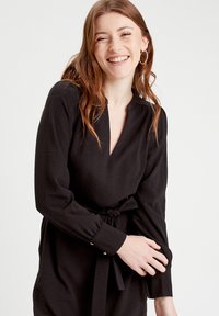 Robe noire portefeuille avec des manches longues, un col en V et une taille à nouer. Elle présente un tissu texturé et des accents de boutons sur les poignets.