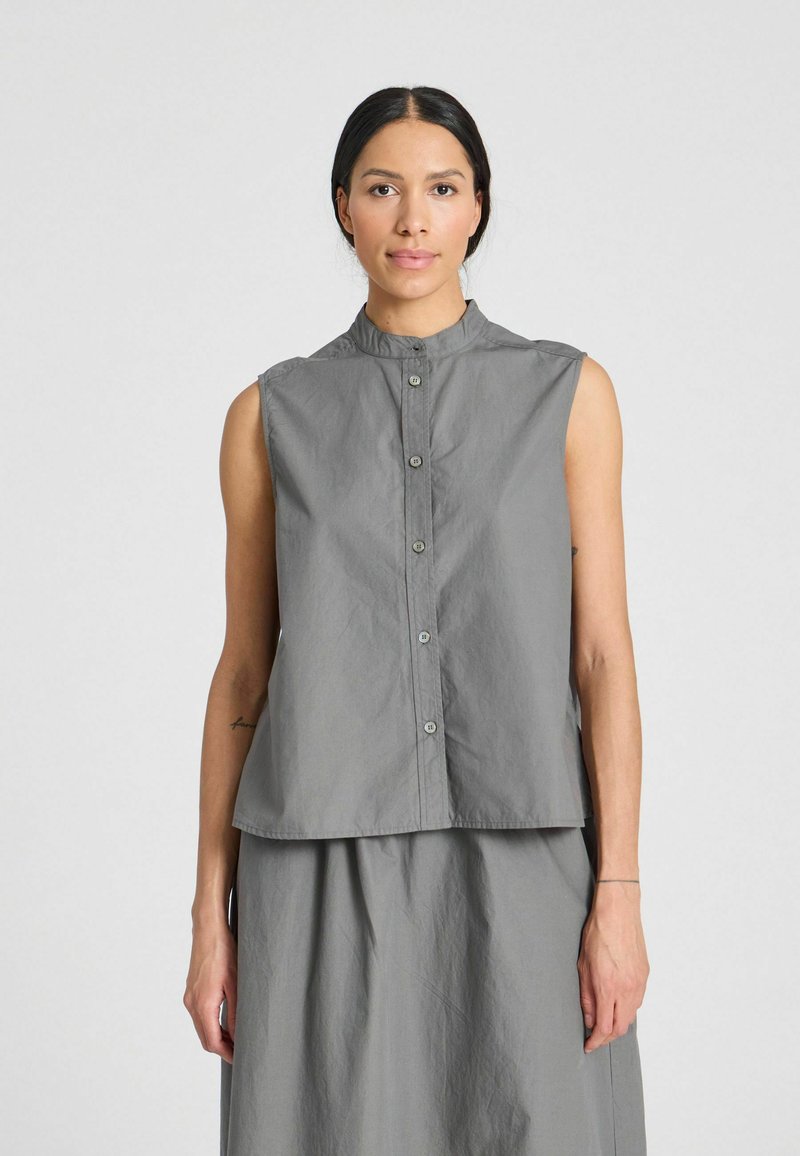 Mouwloze grijze blouse met knopen en een gebande kraag, iets losse pasvorm en subtiele textuur. Gecombineerd met een bijpassende rok voor een samenhangende look.