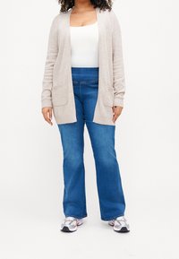 Světlý béžový cardigan s otevřeným předním dílem, žebrovanými manžetami a dvěma předními kapsami, nošený přes bílý top a modré rozšířené džíny. Šedé tenisky doplňují outfit.