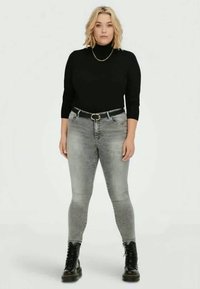 Top de gola alta preto, jeans skinny cinza com um cinto e botas pretas com atacadores. Modelo em pé contra um fundo neutro.