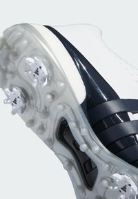 Bílé fotbalové kopačky s námořnickými akcenty, s texturovaným svrškem, průhlednou podrážkou a tvarovanými kolíky pro trakci.