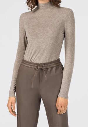 Femme portant un haut moulant à manches longues et col roulé de couleur marron clair ainsi qu'un pantalon marron ample à cordon de serrage, sur un fond uni.