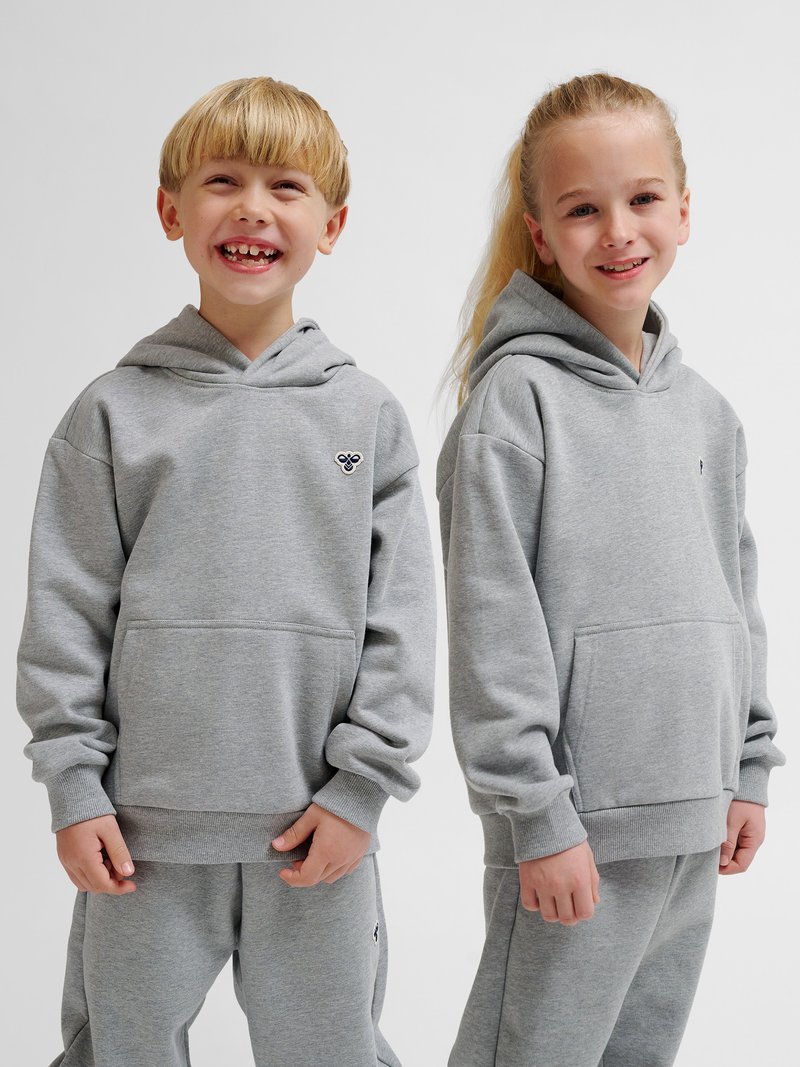 Deux enfants souriants portant des sweats à capuche gris clair assortis et des pantalons, debout côte à côte devant un fond uni.