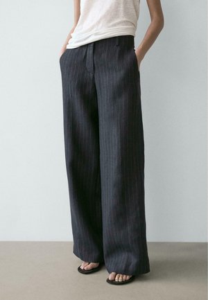 Persona che indossa pantaloni larghi a righe sottili scuri, top chiaro senza maniche e sandali neri con punta aperta, in piedi con le mani nelle tasche.