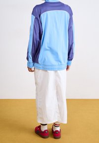 Person med en ljusblå och lila jacka, vita vida byxor, vita strumpor med ränder och röda Puma-sneakers som står på ett senapsgult golv.