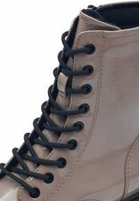 Botín de charol beige con cordones negros, que presenta ojales redondos y una textura lisa y brillante.