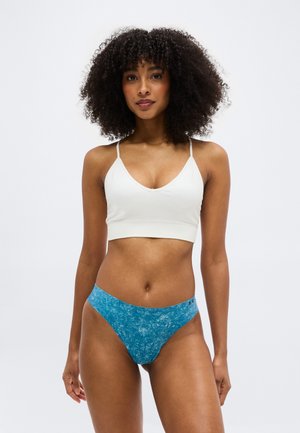 Γυναίκα με σγουρά μαλλιά που φορά λευκό αμάνικο crop top και μπλε υφασμάτινο ψηλοκομμένο εσώρουχο, στέκεται μπροστά σε ένα απλό φωτεινό φόντο.