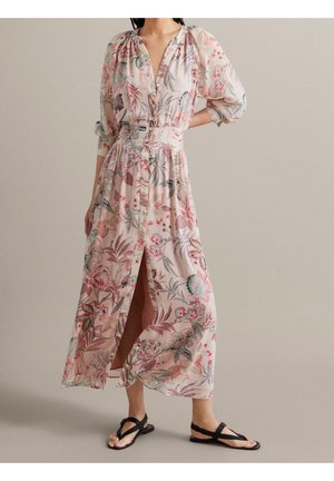 Femme portant une longue robe fleurie rose clair avec des boutons sur le devant et des sandales noires à bout ouvert, debout devant un fond uni.