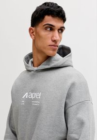 Nuori mies katsoo sivulle, hänellä on yllään vaaleanharmaa huppari, jossa on "aprel"-logo ja rinnassa painetut koordinaatit, neutraali tausta.