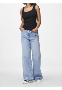 Kvinna i en svart ärmlös knappad top, ljusblå vida jeans med händerna i fickorna, och svartvita sneakers.