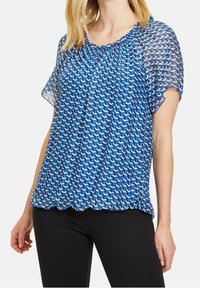 Modrá vzorovaná blůza s krátkými, průsvitnými rukávy. Má nařasený výstřih a zvlněný lem s geometrickým designem v různých odstínech modré.