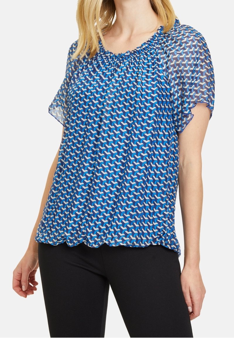 Modrá vzorovaná blůza s krátkými, průsvitnými rukávy. Má nařasený výstřih a zvlněný lem s geometrickým designem v různých odstínech modré.