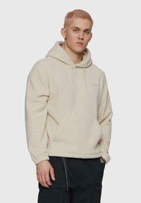 Crème fleece hoodie met een kangoeroezak, trekkoord capuchon en geborduurd logo op de borst. Model draagt donkere cargobroek.