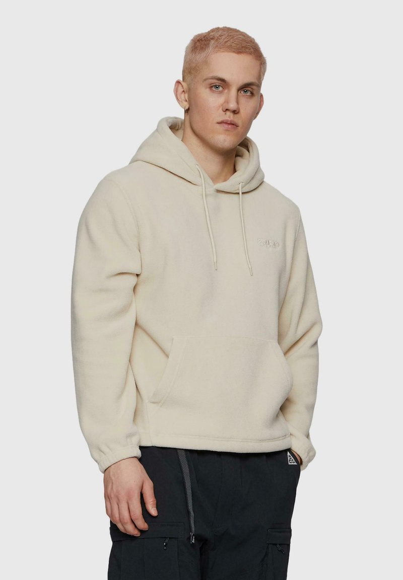 Crème fleece hoodie met een kangoeroezak, trekkoord capuchon en geborduurd logo op de borst. Model draagt donkere cargobroek.