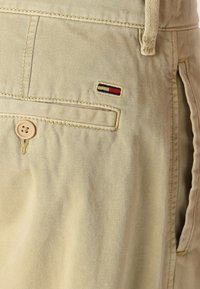 Béžové bavlněné šortky mají zapínání na jeden knoflík, boční kapsu a malou nášivku s červenými, bílými a modrými pruhy na zadní straně.