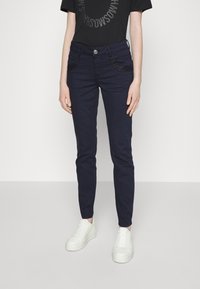 Jeans de mezclilla azul marino con corte slim, con acentos decorativos de bolsillo en negro y cierre de botón. Combinados con zapatillas blancas de suela gruesa.