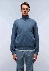 Maglione blu con zip e colletto alto, polsini e orlo a costine. Presenta un piccolo logo sul petto. Abbinato a pantaloni grigi chiari.