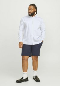 White button-up shirt with long sleeves, paired with navy shorts. Black loafers and white socks with brand lettering. Smooth fabric texture.