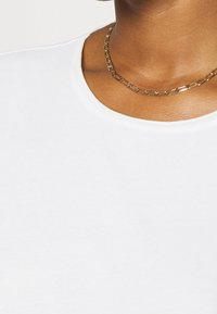 White cotton t-shirt with a round neckline, featuring a smooth texture. A gold chain necklace with alternating links sits at the neck.