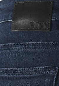 Close-up van een donkerblauwe denim jeans achterzak met een gestikte zwarte leren label waarin "DRYKORN" gebosst is.
