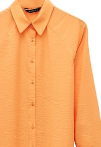 Pomarańczowa koszula z długim rękawem o fakturowanej strukturze, z kołnierzykiem w szpic i guzikami z przodu, rozmiar 35, widoczna od klatki piersiowej wzwyż na białym tle.
