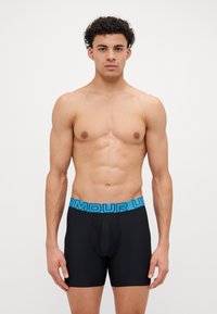 Muskelbepackter Mann steht vor weißem Hintergrund, bekleidet mit schwarzen Boxerbriefs und einem leuchtend blauen Unterwäschebund von Under Armour.