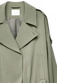 Lysegrøn trenchcoat med brede kraver, skulderrem og rektangulært brystlomme, vist fra øvre bryst til skulderområde.