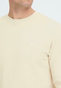 Beżowa koszula z długim rękawem o teksturowanej tkaninie, okrągłym dekolcie i małym logo wytłoczonym po lewej stronie klatki piersiowej.